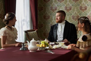 Portrait of classic gentleman enjoying tea with two young ladies sitting at table together in classic ornate room, copy space clipart