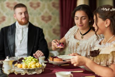 Side view portrait of young gentleman in velvet tailcoat looking at elegant ladies at dining table in classic interior lit by warm lighting clipart