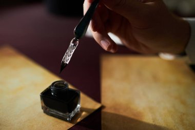 Close up of unrecognizable gentleman dipping pen in ink with focus on transparent glass tip with intricate swirls while writing letters on tinted paper, copy space clipart