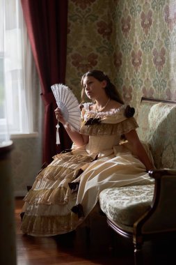 Vertical full length portrait of elegant young lady in classical puffy gown sitting on sofa with fan in hand against ornate wall in vintage interior clipart