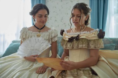 İki genç bayanın birlikte mektup okuduğu ön manzara portresi klasik mavi iç mekan içinde eski koltukta oturmuş parşömenleri okuyorlar.