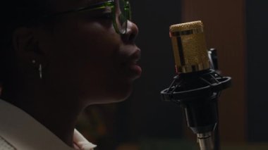 Yakın çekim, gözlüklü Afro-Amerikalı bir kadının mikrofona konuşurken podcast 'i canlı yayın stüdyosunda sıcak ortam aydınlatmasıyla rahat bir atmosfer yaratırken.