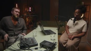 Genç beyaz erkek konuk ve genç zenci kadın sunucu masada mikrofonla konuşuyor, podcast sırasında modern yayın stüdyosunun rahat ortamında sohbet ediyorlar.