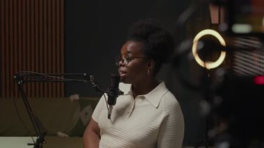 Genç, dost canlısı, siyahi bir radyo sunucusunun kameraya bakıp modern stüdyoda profesyonel yayın ekipmanlarıyla konuşmaya devam ettiği orta boy yakın çekim.