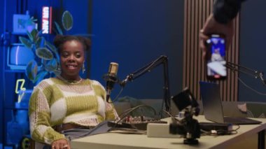 Genç siyahi bir kadının gülümseyen ve kameraya poz veren, mikrofon ve ses ekipmanıyla modern podcast stüdyosunda mavi arka ışıklandırması olan bir adamın telefonundan video kaydediyordu.