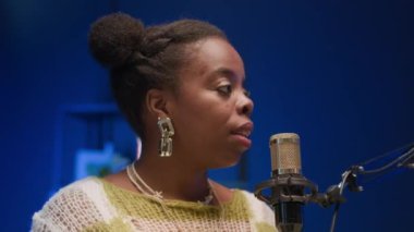 Genç Afrikalı Amerikalı bir kadının parlak küpeler takıp mikrofonun önüne oturduğu, soruları dikkatlice dinlediği ve kayıt stüdyosunda podcast sırasında cevap vermeye hazırlandığı yakın çekim.