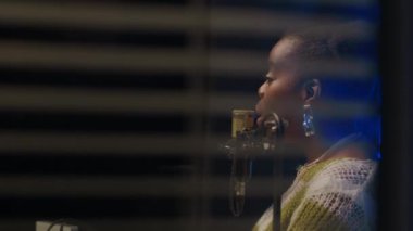 Yayın stüdyosunun cam bölme duvarından görülen, ortam mavisi arka ışıklandırması olan, mikrofon ile konuşan, parlak küpeler takan genç Afrikalı bir kadının yan görüntüsü.