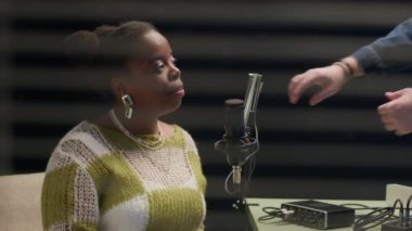 Genç Afrikalı Amerikalı bir kadının kayıt stüdyosunda otururken ve tanınmayan genç bir adamın podcast için ses ekipmanları kurarken ki görüntüsü.