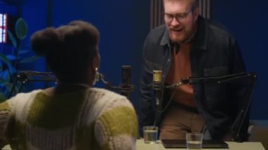 Genç beyaz erkek podcast sunucusu ve genç siyahi bayan konuk teneffüsten sonra masada oturuyorlar, mikrofonla modern stüdyoda çevresel mavi ışıkla rahatça sohbet ediyorlar.