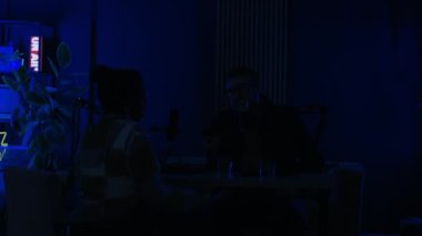 Genç beyaz erkek podcast sunucusu ve mikrofonla konuşan genç siyahi kadın konuk modern kayıt stüdyosunda, karanlıkta neon ışığı parlıyor, sonra ana ışık yanıyor.