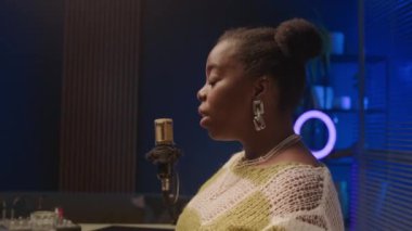 Göğüs hizasında, parlak modern kayıt stüdyosunda cam bölmeli ve mavi arka planda oturan parlak küpeli genç Afro-Amerikan kadını podcast sırasında mikrofonla konuşurken.