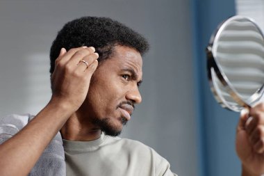 Side view portrait of frowning Black man concerned about hair condition looking at reflection in mirror during grooming routine in bathroom, copy space clipart