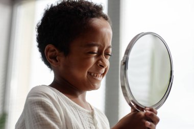 Banyodaki günlük hijyen rutini sırasında küçük bir aynayı tutarken gözleri kapalı gülümseyen mutlu küçük zenci çocuğun portresi.