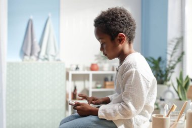 Küçük Afro-Amerikan çocuğun banyoda, ev yaşamında, fotokopi alanında otururken akıllı telefon ekranını dinlerken yan görüntüsü.