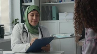 Omuzlarında steteskopla genç ve dost canlısı Müslüman kadın doktorun genç beyaz kadın hastasına sorular sorup açık modern klinikteki ayakta tedavi kartıyla cevaplarını yazışına odaklan.