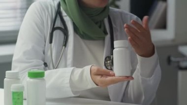 Omuzlarına steteskop takmış, elinde haplı ilaç şişesi olan yeşil başörtüsü takmış ve gündüzleri onları nasıl işe alacağını açıklayan genç bir kadın doktor fotoğrafı.