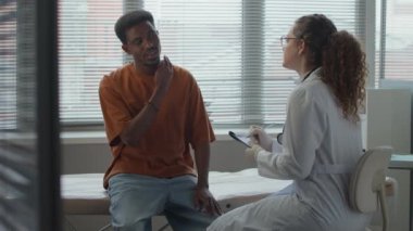 Ağır çekimde, genç siyahi erkek hasta, pencerenin önünde oturuyor. İş yerindeki beyaz kadın doktora boyun sorunları gösteriyor. Dikkatlice dinliyor ve ayakta tedavi kartıyla notlar alıyor.