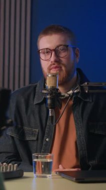 Gözlüklü ve siyah kot ceketli genç beyaz bir adamın dikey orta pozu mikrofon ile konuşarak kayıt stüdyosunda mavi arka plan ışığı ve akustik paneller ile konuşma yapıyor.