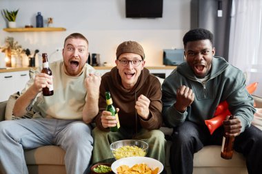 Üç heyecanlı taraftarın portresi evde içki ve atıştırmalık içerken maç zaferini kutluyor.