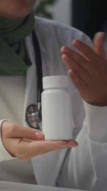 Omuzlarına steteskop takmış, elinde ilaç şişesi olan yeşil başörtüsü takmış ve nasıl içileceğini açıklayan dikey kesilmiş bir kadın doktor fotoğrafı.