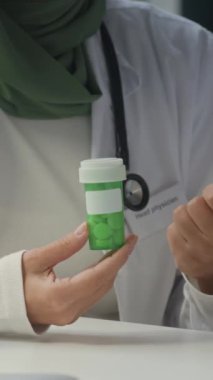 Omuzlarında steteskopla yeşil başörtüsü ve beyaz önlüklü yeşil ilaç şişesinde ilaç yazan genç kadın doktorların dikey, yavaş çekimleri.
