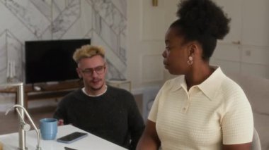 Genç Afrikalı Amerikalı kadın mutfak tezgahında dizüstü bilgisayar başında çalışıyor ve tekerlekli sandalyedeki genç beyaz adama kupa veriyor, ona yaklaşıyor, modern evin rahat atmosferinde sohbet ediyorlar.