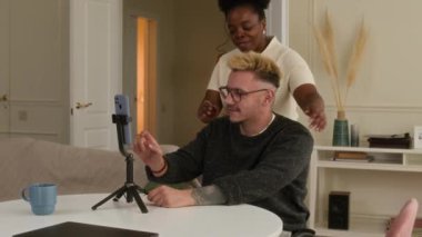 Tekerlekli sandalyedeki genç beyaz adam, rahat bir evde, telefonla video bloğu kaydediyor. Genç siyahi bir kadın ona sarılıyor, kameraya el sallıyor ve köpek de onlara katılıyor.