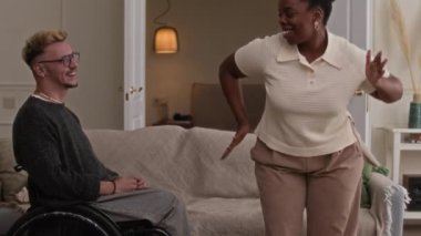 Genç, neşeli siyahi kadın, robot dansı hareketlerini tekerlekli sandalyedeki genç beyaz adama gösteriyor. Arkasından tekrarlıyor, hem gülüyor, hem de modern evde eğleniyor.