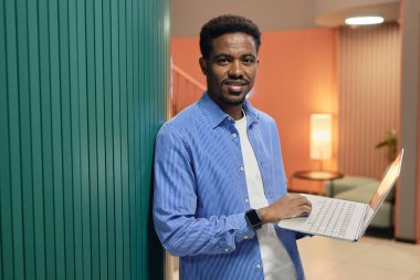 Ofis için mavi gömlekli, elinde dizüstü bilgisayarla iş yerinde duvara yaslanmış gülümseyen Afro-Amerikan erkek çalışanın orta boy portresi.