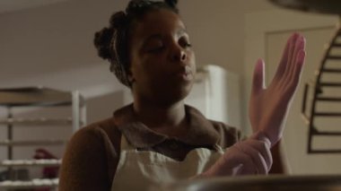 Siyah bir kadının apronlu yavaş çekimleri, sıcak mutfakta eldivenlerini dikkatlice takması, hijyen ve hamur işi üretim sürecine olan bağlılığını vurgulaması,