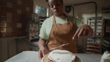 Tilt down shot of skilled African American female pastry chef spreading white cream over layered cake on rotating stand