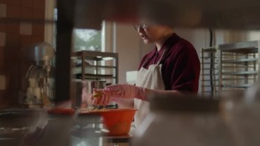 Genç fırıncının gözlüklü, pembe eldivenli plastik kasede hamur işi şekillendirirken modern, iyi donanımlı mutfakta paslanmaz yemek masasının yanında durduğu orta yavaş bir fotoğraf.