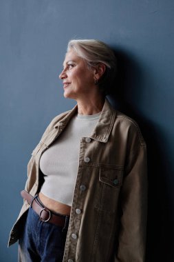 Vertical side view portrait of modern elegant senior woman smiling while posing relaxed against blue wall in studio with natural lighting accentuating profile clipart