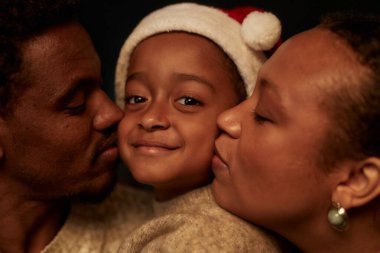 Close up of mother and father kissing little boy on both cheeks with child wearing Santa hat and smiling at camera clipart