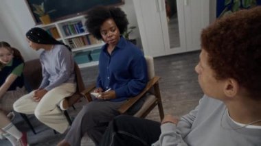 Genç siyahi kadın psikolojisi uzmanının grup terapisi sırasında çift ırklı bir çocukla modern okul ofisindeki çok ırklı z çocuklarıyla aynı çemberde otururken yüksek açılı görüntüsü.