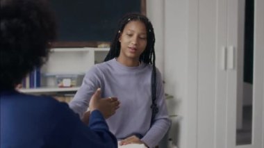 Orta ölçekli, örgülü saçlı Afrikalı Amerikalı bir genç kızın önünde oturan tanınmayan bir kadınla el hareketleriyle selamlama hareketi yaptığı bir fotoğraf.