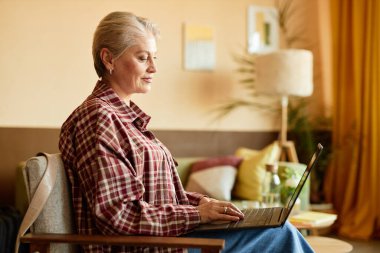 İnternette çalışırken, iş yerinde ya da ev ofisinde rahat koltukta oturan, rahat tişörtlü, gülümseyen olgun bir kadının yan görüntüsü, boşluğu kopyala