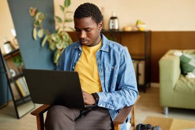 Genç Afrikalı Amerikalı yazılım geliştiricisinin evdeki ya da işyerindeki rahat koltukta otururken dizüstü bilgisayara kod yazarken orta boy görüntüsü.