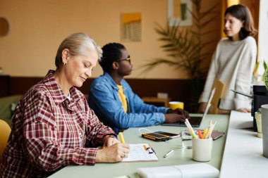 Side view of mature female employee writing in calendar table planning monthly tasks while sitting at working desk in office, copy space clipart