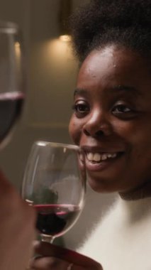 Dikey yakın plan çekimde, keyifli, Afro-Amerikalı bir kadın, rahat bir kazak giyiyor, kırmızı şarap kadehleriyle kadeh kaldırıyor, tanınmayan biriyle sakin romantik bir akşam geçiriyor.