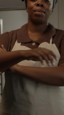 Dikey eğim, önlüklü genç siyahi kadın pasta şefinin orta yavaş portresi. Elleri katlı ve kameraya gülümsüyor, arka planda paslanmaz çelik askıyla mutfakta duruyor.