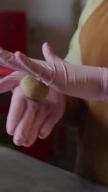 Dikey olarak, tanınmayan Afro-Amerikan fırıncının pembe eldivenli yavaş çekimleri mutfakta tezgahın üzerinde küçük bir hamur topunu dikkatle yuvarlıyor.