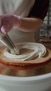 Pembe eldivenli tanınmayan fırıncının dikey yavaş çekimleri. Pastanın üzerine beyaz krema serpiştiriyor. İyi donanımlı mutfak ortamında pikap çantasıyla duruyor.