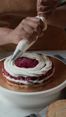 Önlüklü tanınmayan kadın fırıncının dikey ve yavaş çekimleri ahudududulu kekin üzerine boru torbasıyla tatlı beyaz krema yayıyor.