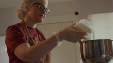 Düşük açılı bel üstü, tek kullanımlık kepli, eldivenli ve gözlüklü beyaz kadın fırın işçisinin profesyonel mutfak ortamında hamur işi malzemelerini hazırlarken karıştırma kabına un dökmesi.