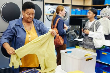 Medium shot of senior African American woman folding shirt made of delicate fabric at table checking results of washing in public laundry room with people chatting in background, copy space clipart