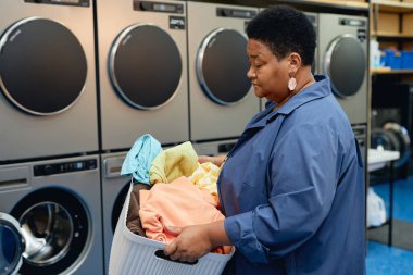 Side view of senior African American woman carrying basket of laundry preparing colored clothes for washing at self service laundromat, copy space clipart