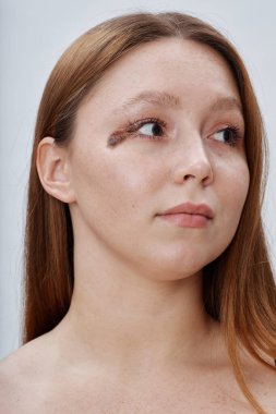 Yüzünde doğum lekesi olan güzel kızıl saçlı kızın dikey portresi beyaz arka planda poz verirken sakin ve kendinden emin bir ifadeyle başka tarafa bakıyor.