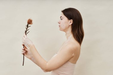 Stüdyoda bej arkaplanda poz veren kırılgan çiçekleri tutan, Vitiligo 'lu huzurlu genç bir kadın. Benzersiz deri desenli doğal güzelliği kucaklayan, kopyalama alanı.