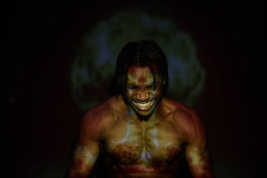 Gömleksiz siyah adamın göğüs portresi öfkeyle bağırıyor dişlerini gösterirken projektörde siyah arka planda üstsüz vücudunda nükleer patlama görüntüsü var.